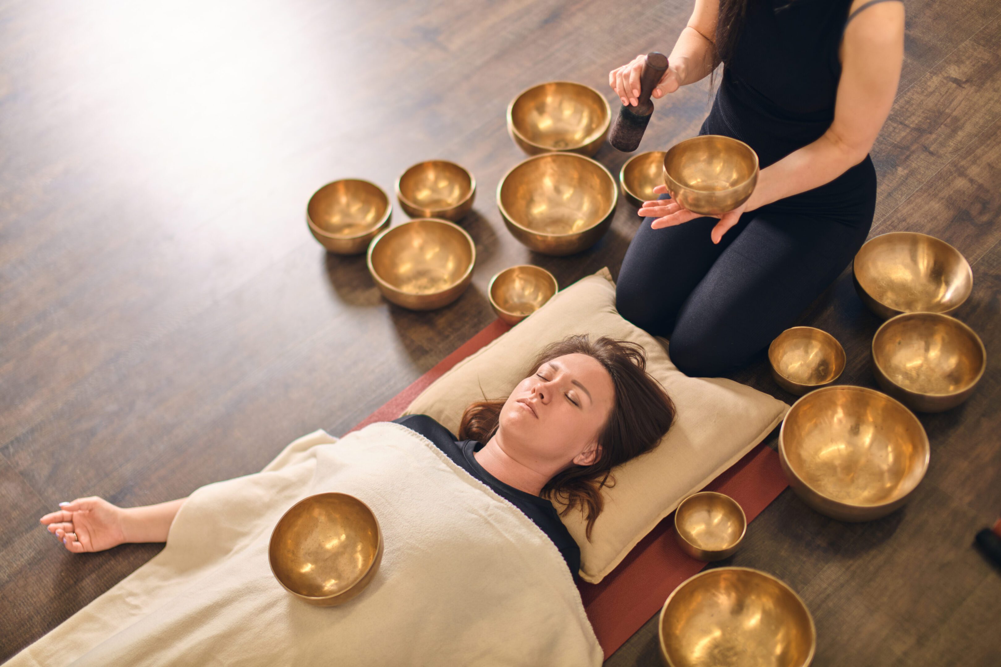 Young woman lies relaxed, surrounded by singing bowls, in serene setting. Singing bowl rests on her torso, creating harmonious ambiance. Soft lighting enhances tranquility