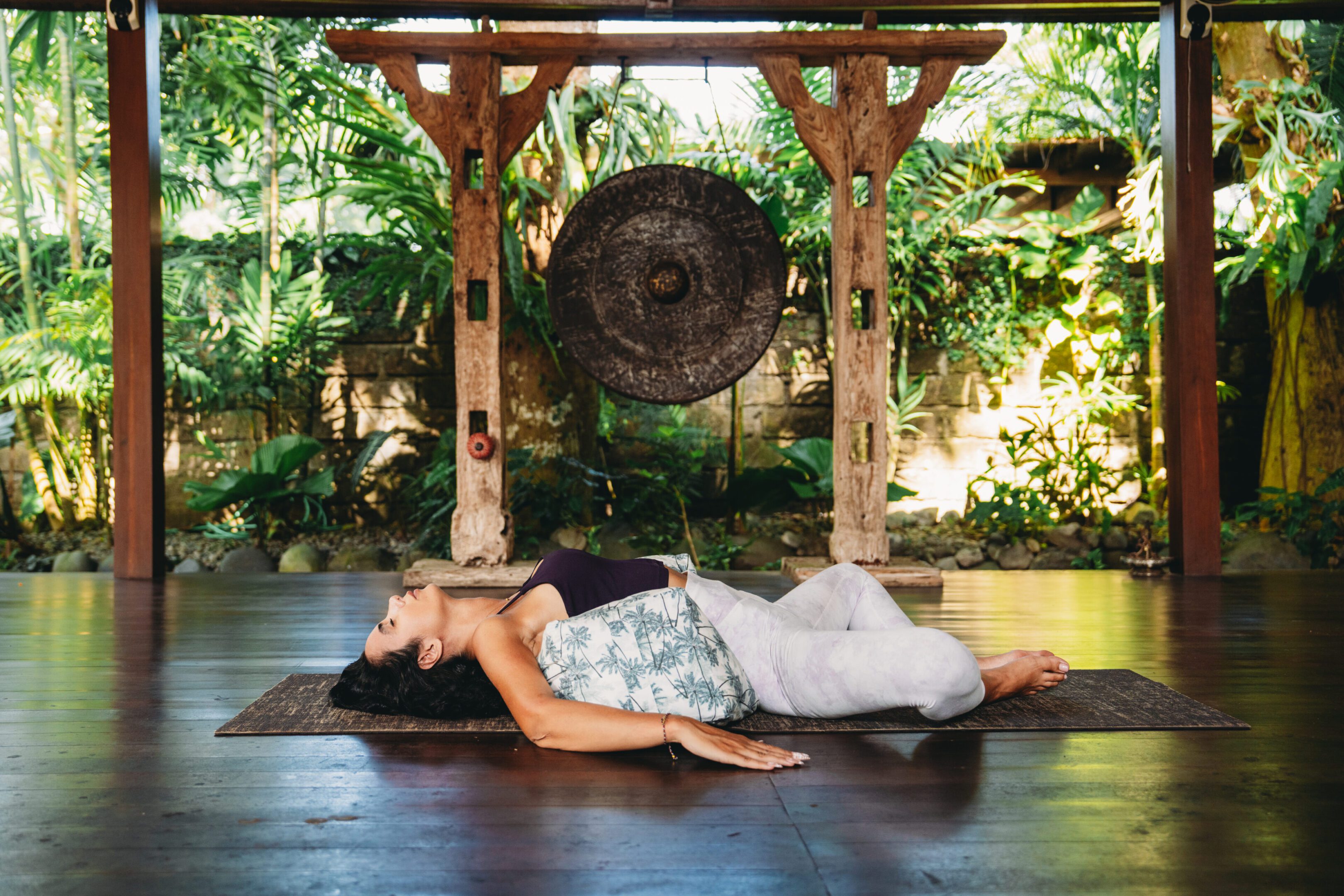 Woman lying on a yoga mat with a cushion, relaxing during a sound bath session with a large gong in a tropical setting