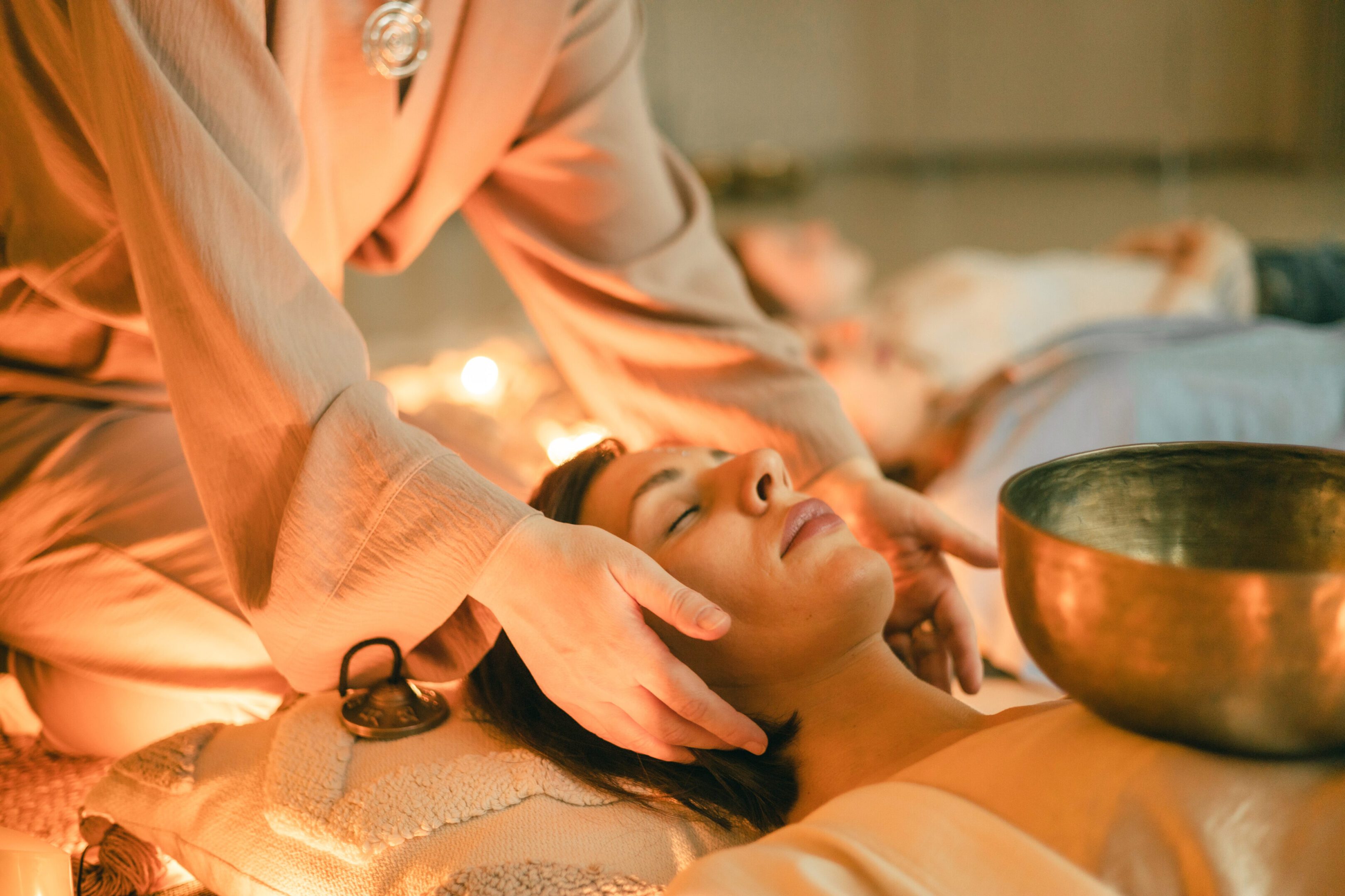 A therapist gently supports a woman’s head during a sound healing session with a Tibetan bowl. Warm lights, soft textures, and calm energy create a peaceful environment for deep relaxation and mindfulness.
