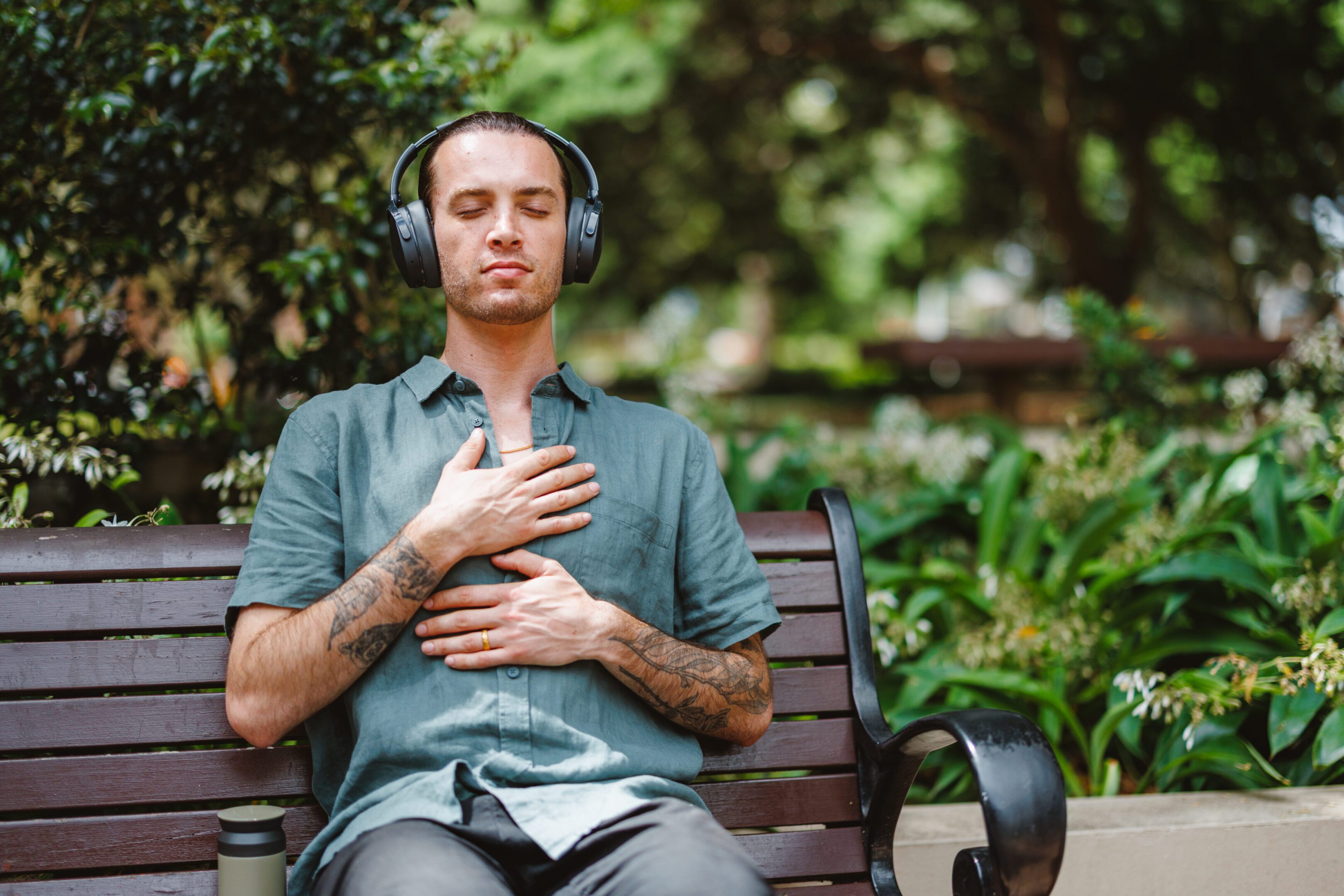 A mid adult Caucasian non-binary person sits calmly on a park bench in Sydney, listening to guided meditation with a relaxed expression. Surrounded by lush greenery, they appear content and at peace, fully immersed in the serene outdoor setting.